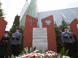 Widok na pomnik od dołu, widać wiązanki kwiatów i asystę policyjną.