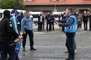 Mężczyzna z zespołu antykonfliktowego w niebieskiej kamizelce wyciąga ręce w stronę grupy mężczyzn przebranych za kibiców.