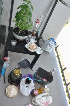 Zdjęcie z góry na posadzkę balkonowa, na której stoją słoiki w reklamówkach, wiadro, szyfelka, krzew marihuany w dniczce na stole.