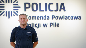 Policjant w granatowym mundurze stoi bez czapki a w tle widnieje logo Komedny Powiatowej Policji w Pile