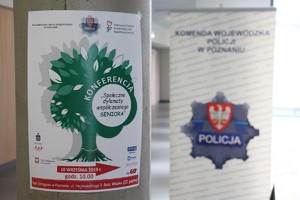Plakat konferencji senioralnej wisi na słupie, a za nim stoi baner KWP w Poznaniu