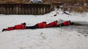 Pokaz ratownictwa wodnego na lodzie. Jeden z policjantów w stroju nurka jest w przeręblu, a trzech leży na lodzie i próbują go wyciągnąć.